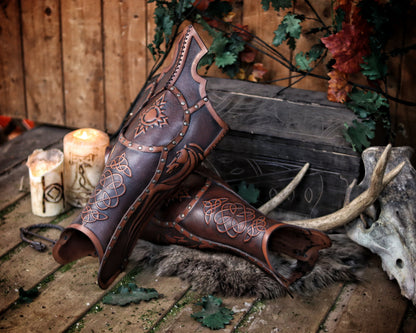 Side view of two Celtic leather greaves placed on fur, next to lit candles, antlers, and a carved chest. The embossed details—sun symbol, horse, and knotwork—stand out under warm lighting. The greaves are slightly tilted, showing the curve of the shin and opening at the calf. The scene recreates a fantasy camp with medieval atmosphere, perfect for showcasing handcrafted Viking or Celtic LARP leg armor made with aged vegetable-tanned leather.