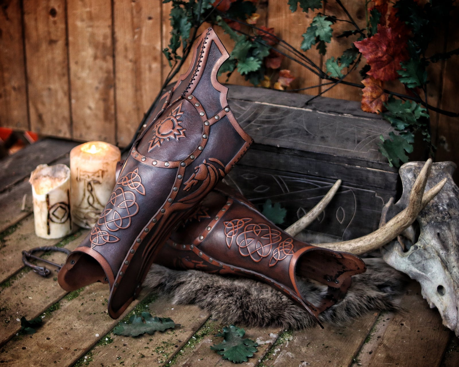 Side view of two Celtic leather greaves placed on fur, next to lit candles, antlers, and a carved chest. The embossed details—sun symbol, horse, and knotwork—stand out under warm lighting. The greaves are slightly tilted, showing the curve of the shin and opening at the calf. The scene recreates a fantasy camp with medieval atmosphere, perfect for showcasing handcrafted Viking or Celtic LARP leg armor made with aged vegetable-tanned leather.