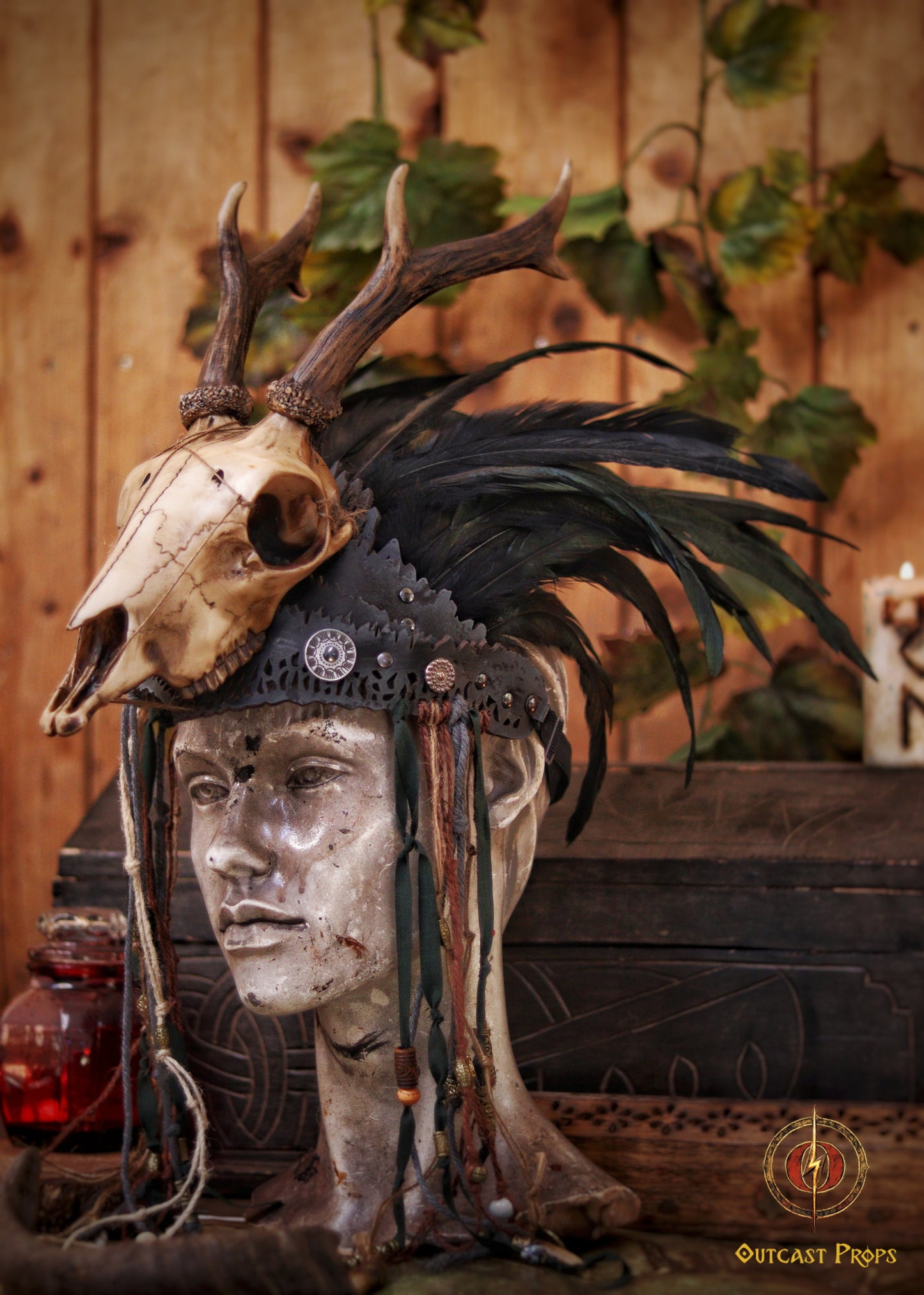A Celtic adorned tiara with a resin skull, deer horns, feathers, bone pieces, beads, and fangs displayed on a table with a wooden background and candles. Beltane Headpiece tribal deer horns skull and feathers tiara leather and resin