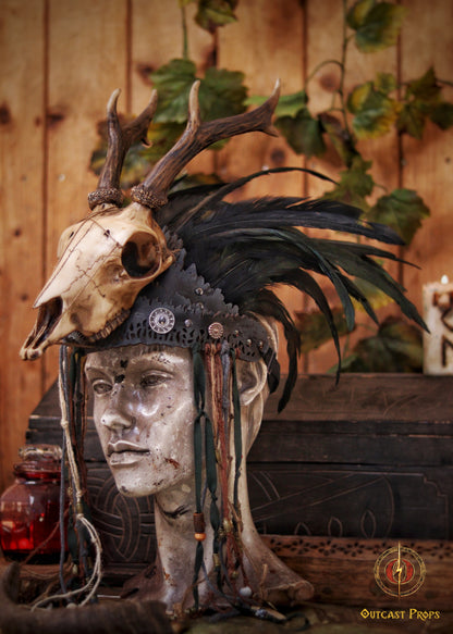 A Celtic adorned tiara with a resin skull, deer horns, feathers, bone pieces, beads, and fangs displayed on a table with a wooden background and candles. Beltane Headpiece tribal deer horns skull and feathers tiara leather and resin