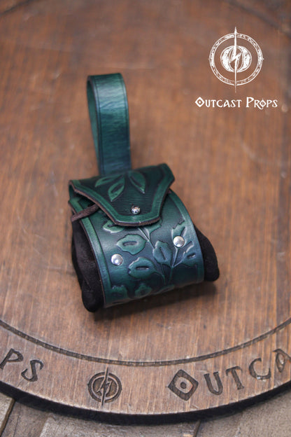A green leather coin bag with an embossed floral design rests on a wooden surface with the Outcast Props logo above it. The pouch combines a structured vegetable tanned leather flap with soft black suede sides, creating an elegant elven accessory ideal for renfaire outfits, herbalist costumes, witches or nobles. The rounded shape and belt loop make it a practical leather coin bag or fantasy dice pouch for LARP and daily carry.