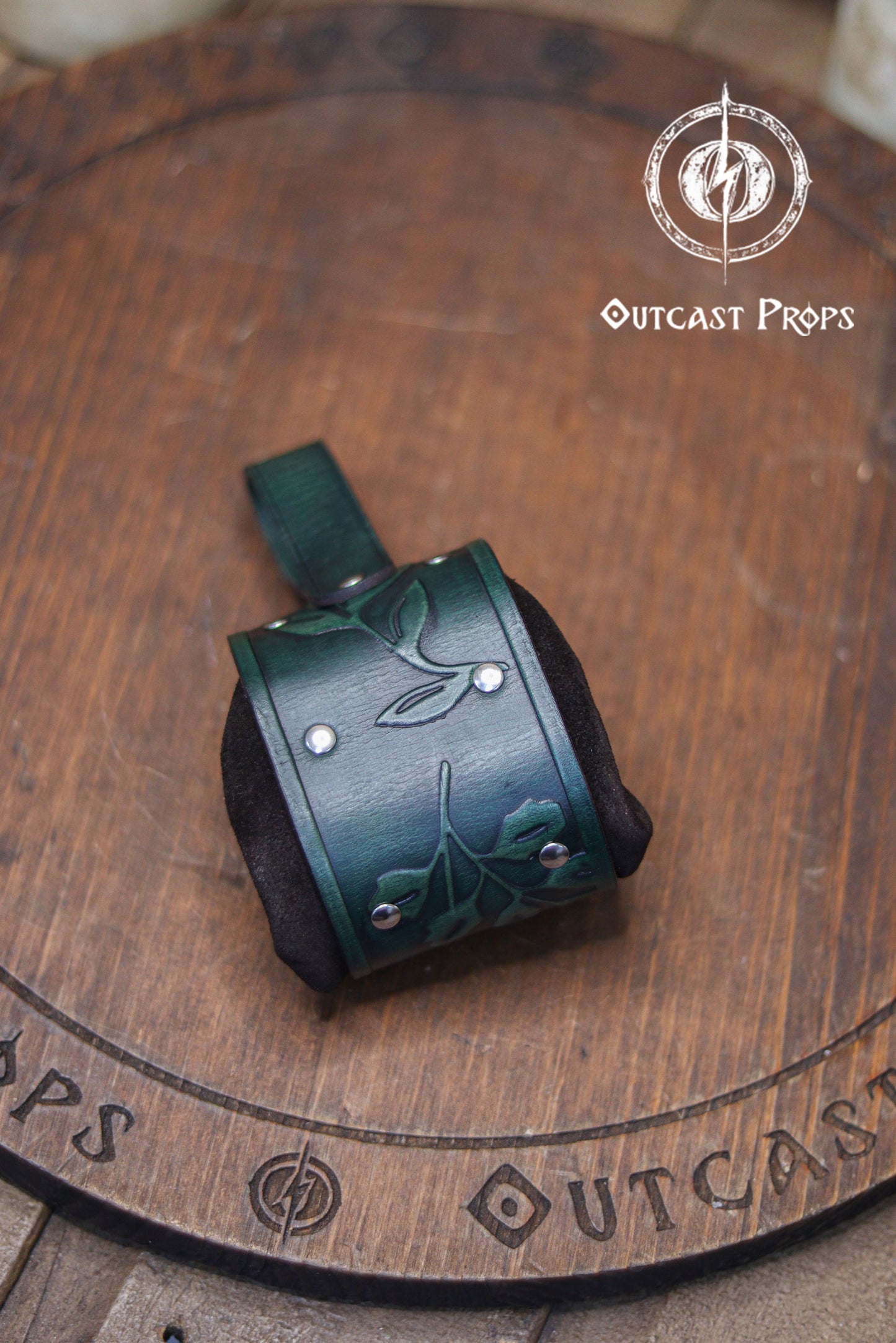 Close up of the back of a green embossed leather coin bag displayed on a round wooden board engraved with the Outcast Props logo. The pouch features botanical leaf motifs, silver rivets and soft suede sides. Designed as an elven herbalist bag, it attaches to the belt and works perfectly for carrying coins, keys or dice at a renaissance faire, renfaire or LARP event. Its handcrafted look and floral pattern make it a refined fantasy dice pouch.