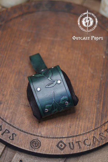 Close up of the back of a green embossed leather coin bag displayed on a round wooden board engraved with the Outcast Props logo. The pouch features botanical leaf motifs, silver rivets and soft suede sides. Designed as an elven herbalist bag, it attaches to the belt and works perfectly for carrying coins, keys or dice at a renaissance faire, renfaire or LARP event. Its handcrafted look and floral pattern make it a refined fantasy dice pouch.
