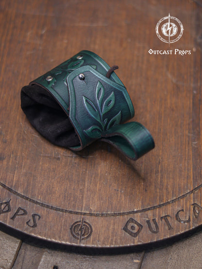 Angle view of a green floral embossed leather coin pouch lying on a wooden surface marked with the Outcast Props emblem. The flap shows elegant leaf carving, while the soft suede base forms a rounded pocket. This renfaire pouch is ideal for elves, druids, witches or herbalists and carries coins, dice or tiny items securely on the belt. A perfect fantasy accessory for renaissance faire costumes and LARP characters seeking natural themed details.