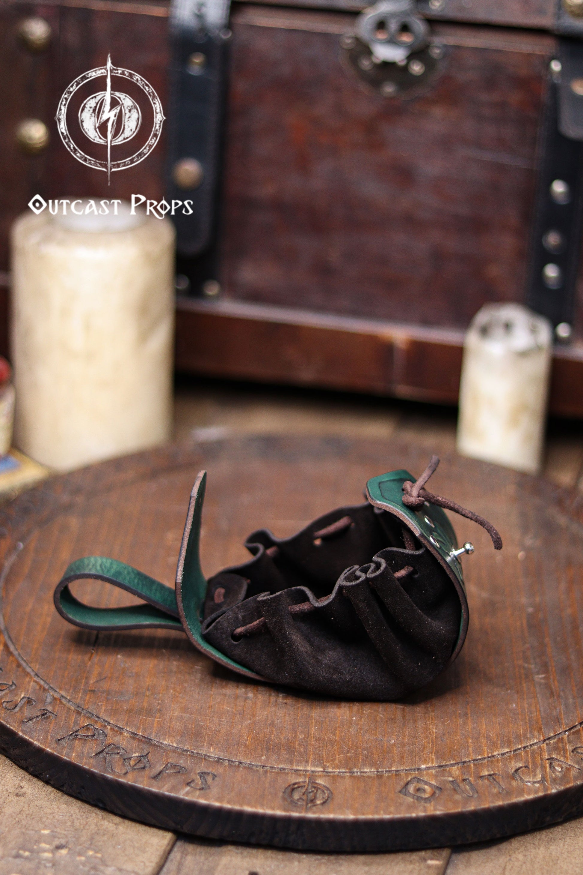 A green leather coin pouch is shown open on a wooden surface, revealing its soft dark suede interior with gathered folds and a drawstring closure. The structured leather flap features botanical embossing, while the back loop allows it to hang from a belt. In the blurred background there is a wooden chest and candles, creating a fantasy workshop atmosphere. This elven herbalist renfaire pouch is ideal for coins, dice or small items at a renaissance faire or LARP.
