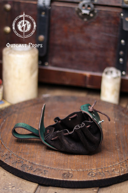 A green leather coin pouch is shown open on a wooden surface, revealing its soft dark suede interior with gathered folds and a drawstring closure. The structured leather flap features botanical embossing, while the back loop allows it to hang from a belt. In the blurred background there is a wooden chest and candles, creating a fantasy workshop atmosphere. This elven herbalist renfaire pouch is ideal for coins, dice or small items at a renaissance faire or LARP.