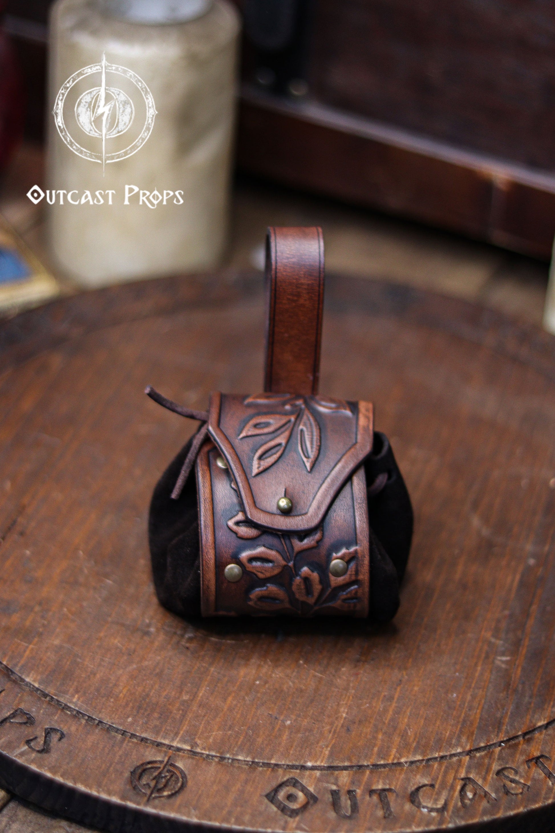 A brown leather coin pouch with an embossed floral pattern stands centered on a round wooden board engraved with the Outcast Props logo. The soft suede sides create a rounded shape, while the looped strap allows it to hang from a belt. Warm brown tones and botanical carvings give it an elegant elven or herbalist look, ideal for witches, nobles and renfaire characters. Perfect as a leather coin bag or fantasy dice pouch for LARP, cosplay or daily use.