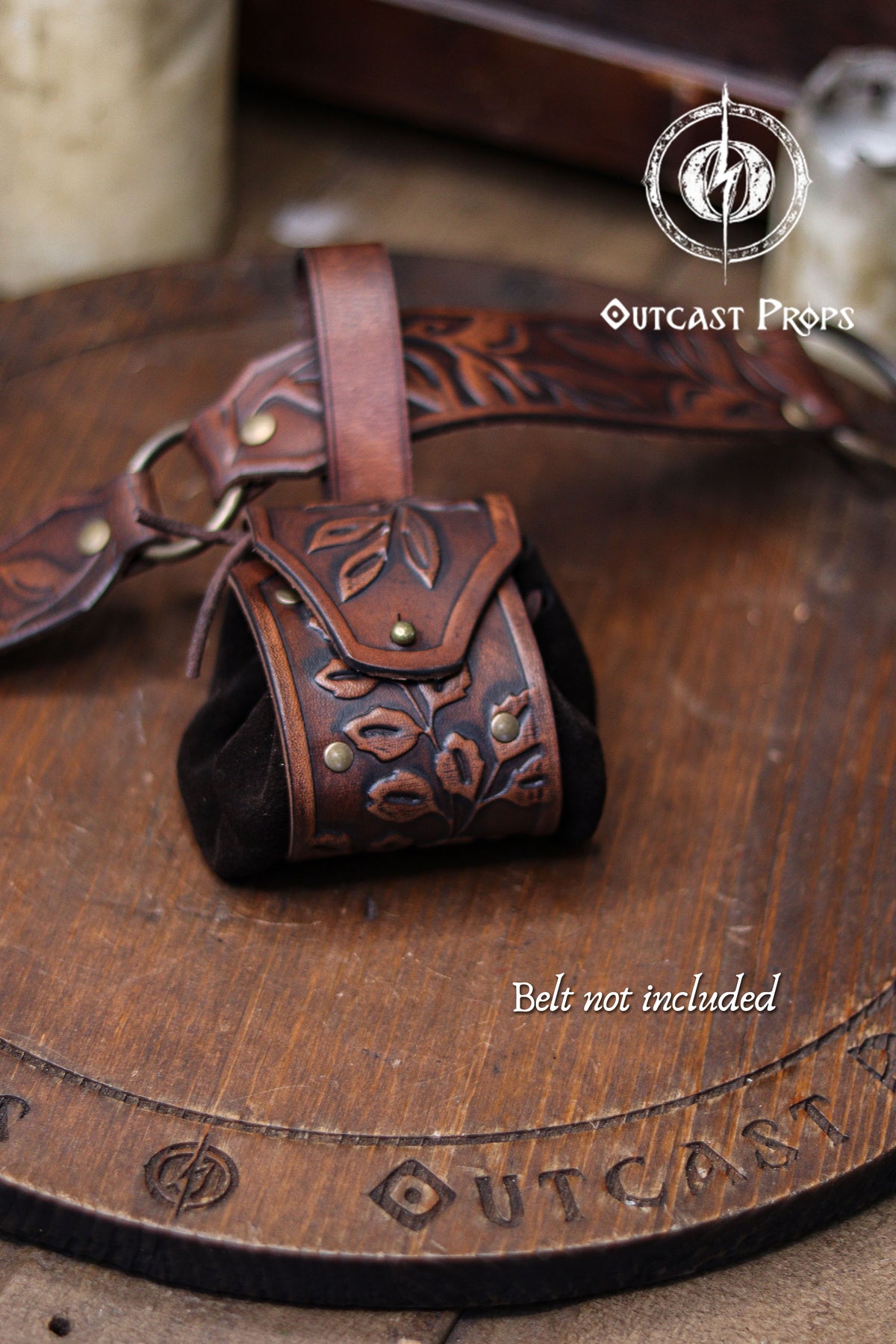 A brown floral embossed leather coin pouch hangs from a matching tooled belt resting on a wooden board. Text reading Belt not included appears at the bottom. The pouch features brass rivets, a suede body and a botanical flap, making it suitable for elven nobles, herbalists or witches at a renaissance faire or renfaire. Its handcrafted style and warm aged finish create a fantasy coin bag ideal for holding dice, keys or small essentials.
