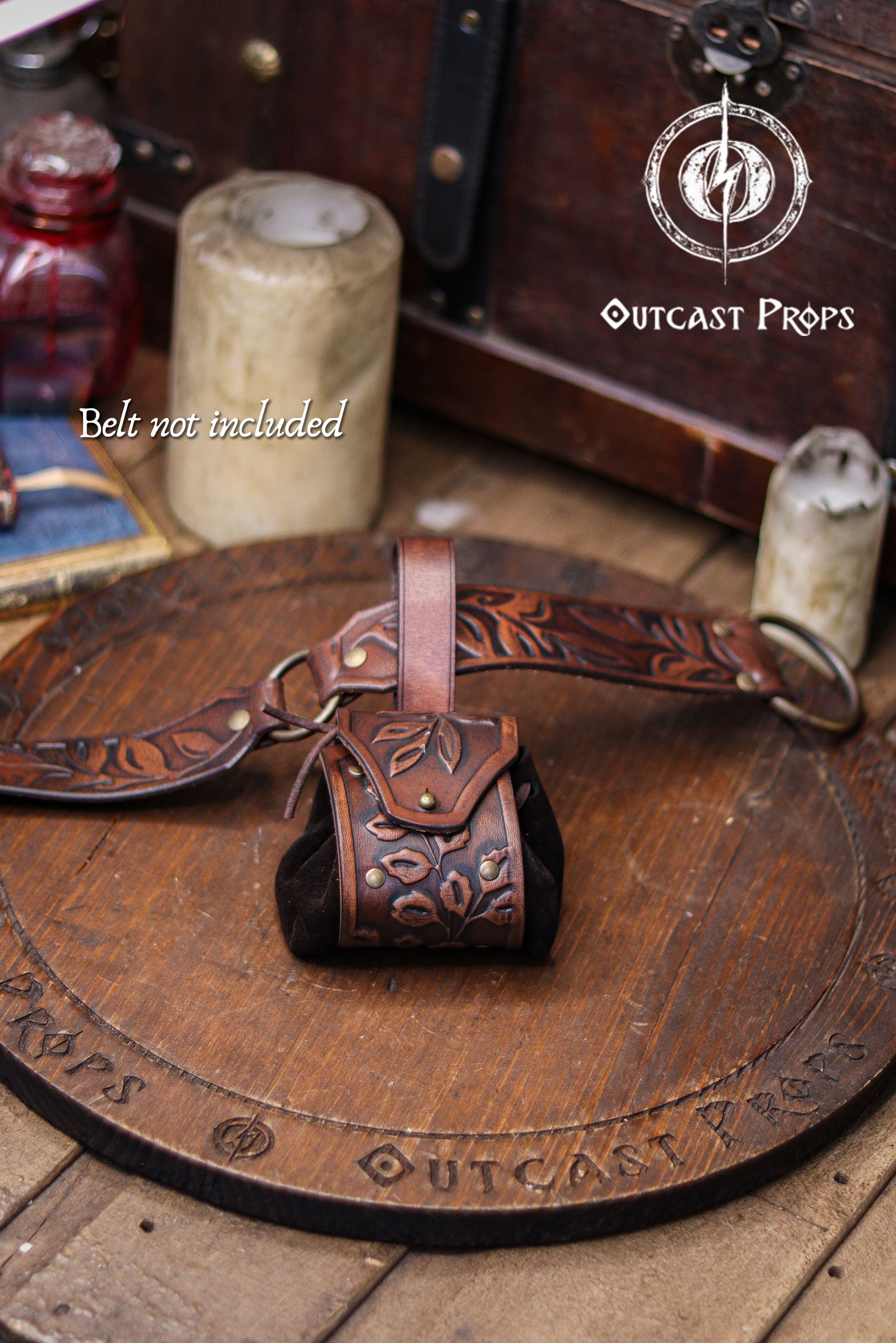 Brown leather belt with pouch on a wooden surface, featuring 'Outcast Props' branding. A brown floral embossed leather coin pouch hangs from a matching tooled belt resting on a wooden board. Text reading Belt not included appears at the bottom. The pouch features brass rivets, a suede body and a botanical flap, making it suitable for elven nobles, herbalists or witches at a renaissance faire or renfaire. Its handcrafted style and warm aged finish create a fantasy coin bag ideal for holding dice, keys or sma
