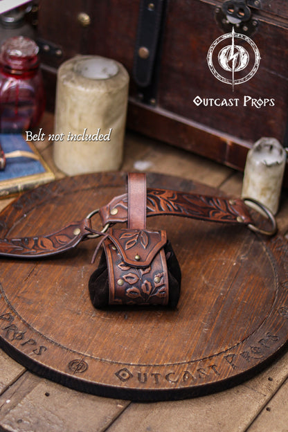 Brown leather belt with pouch on a wooden surface, featuring 'Outcast Props' branding. A brown floral embossed leather coin pouch hangs from a matching tooled belt resting on a wooden board. Text reading Belt not included appears at the bottom. The pouch features brass rivets, a suede body and a botanical flap, making it suitable for elven nobles, herbalists or witches at a renaissance faire or renfaire. Its handcrafted style and warm aged finish create a fantasy coin bag ideal for holding dice, keys or sma