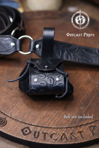A black floral embossed leather coin pouch sits next to a matching embossed belt on a wooden surface. Text Belt not included appears at the bottom. The pouch mixes suede and vegetable tanned leather, creating a dark refined accessory for witches, nobles, druids or elven characters. Ideal for renfaire or renaissance faire costumes, it serves as a fantasy dice pouch or small coin bag for LARP, cosplay or everyday carry.