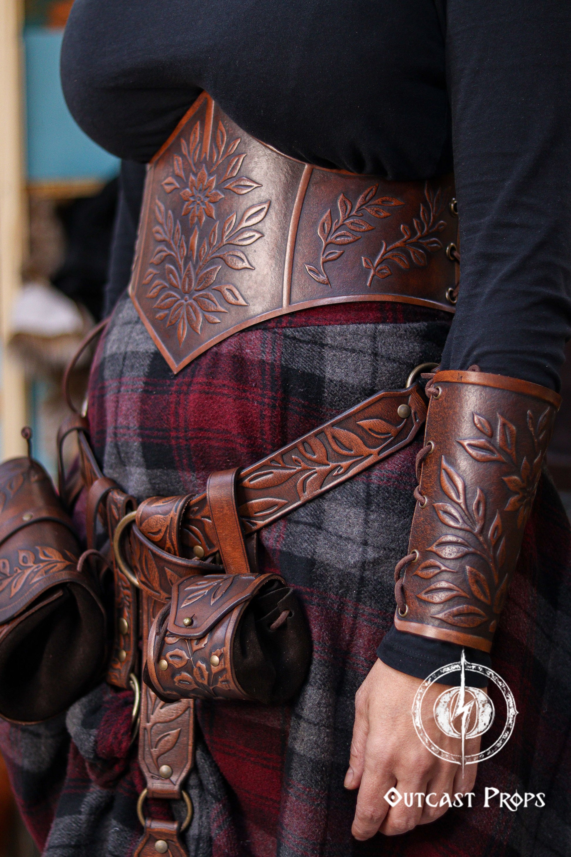 Side view of a model wearing an aged brown floral leather underbust corset belt paired with matching accessories from the same floral set. The embossed leaves and artisan aging give it a rich renaissance faire aesthetic. The corset cinches the waist beautifully, perfect for elven characters, witches, herbalists, fantasy cosplay and LARP costumes. The belt pouches and skirt hike add practical medieval flair.