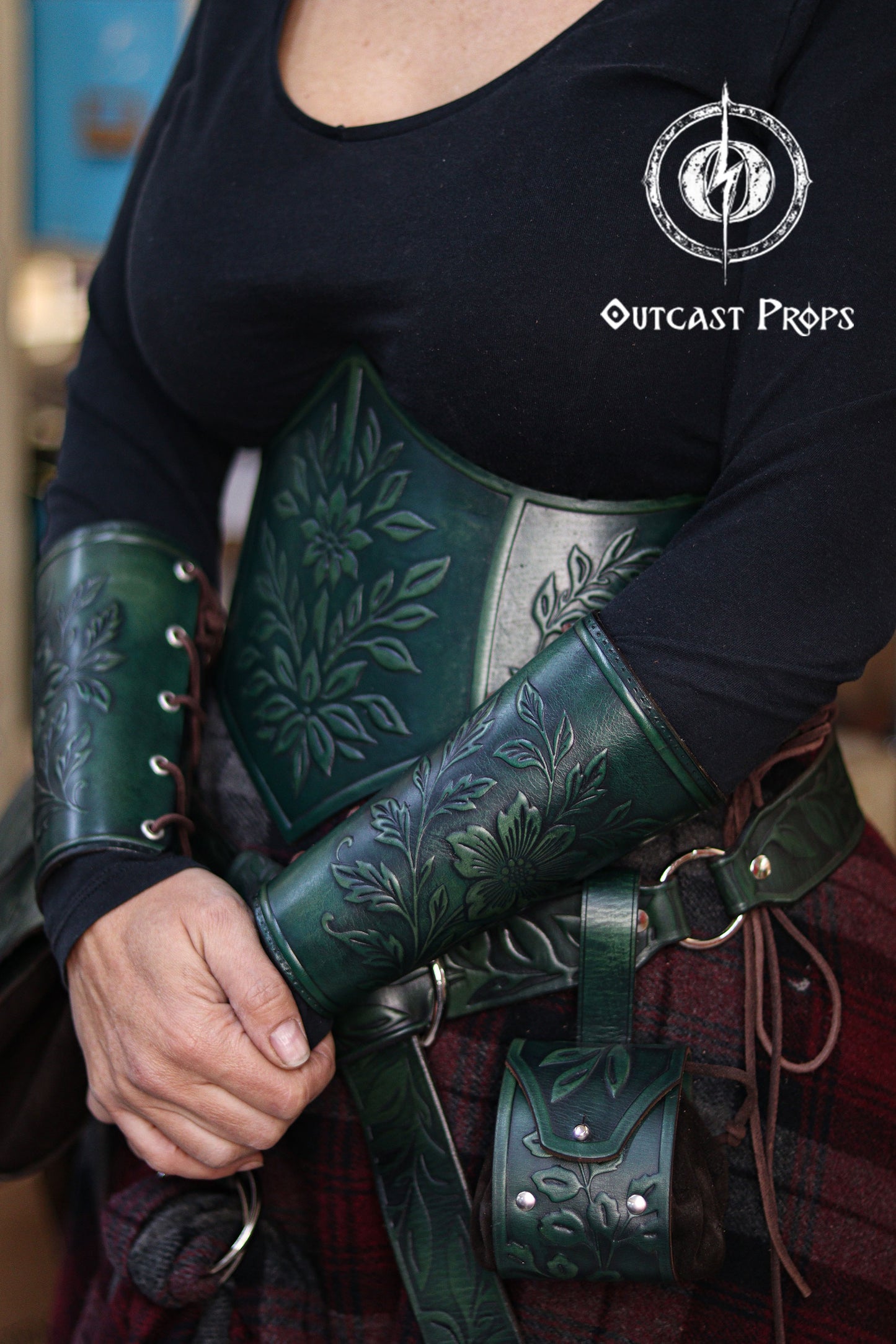 The green floral leather underbust corset belt is shown together with the matching bracers, creating a cohesive renaissance faire outfit. The embossed flowers and leaves evoke an elven forest theme, ideal for herbalist characters, witchcore fashion, fantasy cosplay and LARP costumes. The rich green dyes, side lacing and handcrafted aging techniques highlight the artisan quality of the full floral set by Outcast Props.
