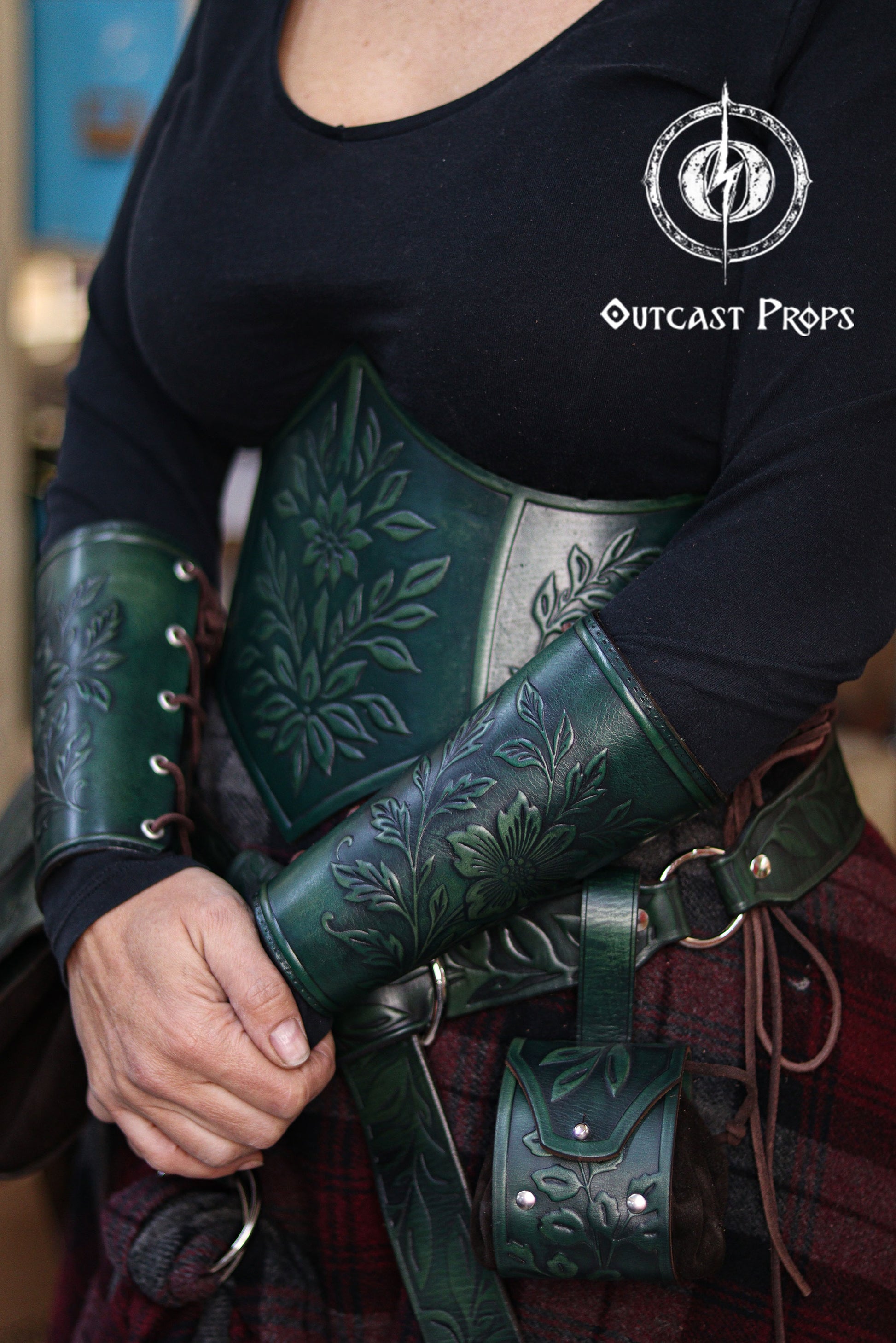 The green floral leather underbust corset belt is shown together with the matching bracers, creating a cohesive renaissance faire outfit. The embossed flowers and leaves evoke an elven forest theme, ideal for herbalist characters, witchcore fashion, fantasy cosplay and LARP costumes. The rich green dyes, side lacing and handcrafted aging techniques highlight the artisan quality of the full floral set by Outcast Props.