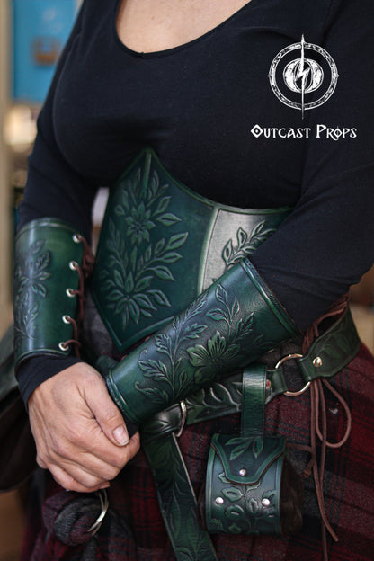 The green floral leather underbust corset belt is shown together with the matching bracers, creating a cohesive renaissance faire outfit. The embossed flowers and leaves evoke an elven forest theme, ideal for herbalist characters, witchcore fashion, fantasy cosplay and LARP costumes. The rich green dyes, side lacing and handcrafted aging techniques highlight the artisan quality of the full floral set by Outcast Props.