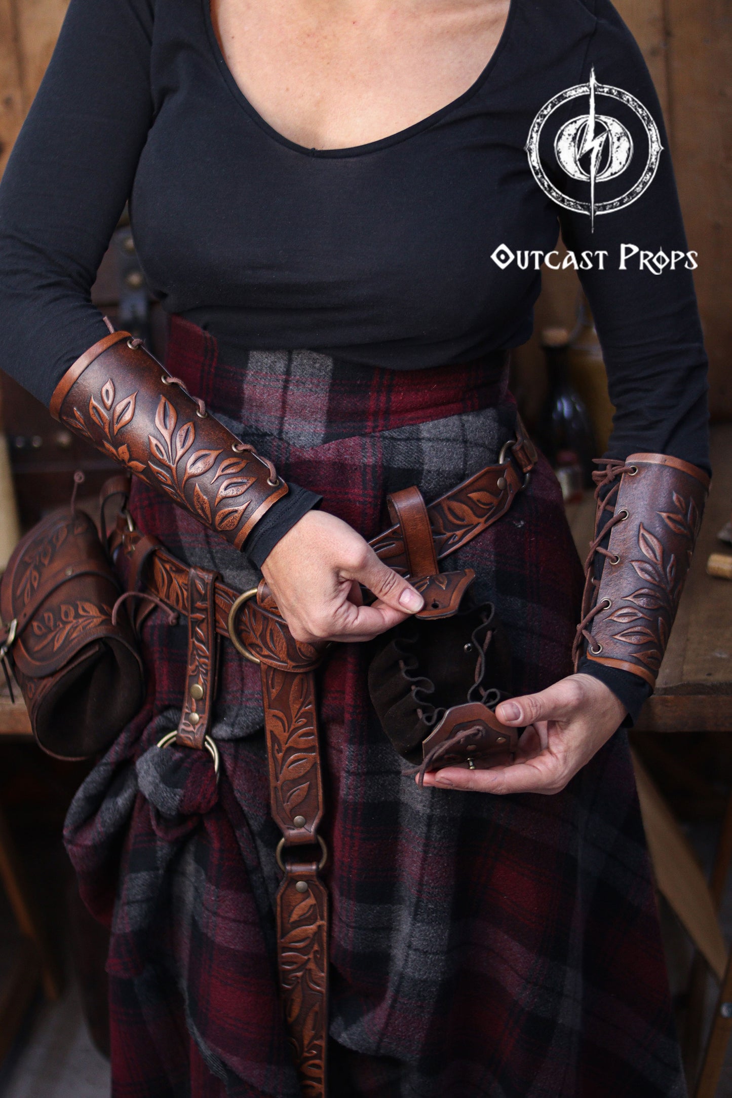A person wearing brown embossed leather bracers and a matching belt demonstrates how to open a floral leather coin pouch. The plaid skirt and rustic workshop background create a renaissance faire atmosphere. The pouch’s suede interior and drawstring are visible as it is held open. This handcrafted renfaire pouch suits elves, witches, herbalists and nobles, working as a leather coin bag or fantasy dice pouch for immersive LARP and cosplay costumes.