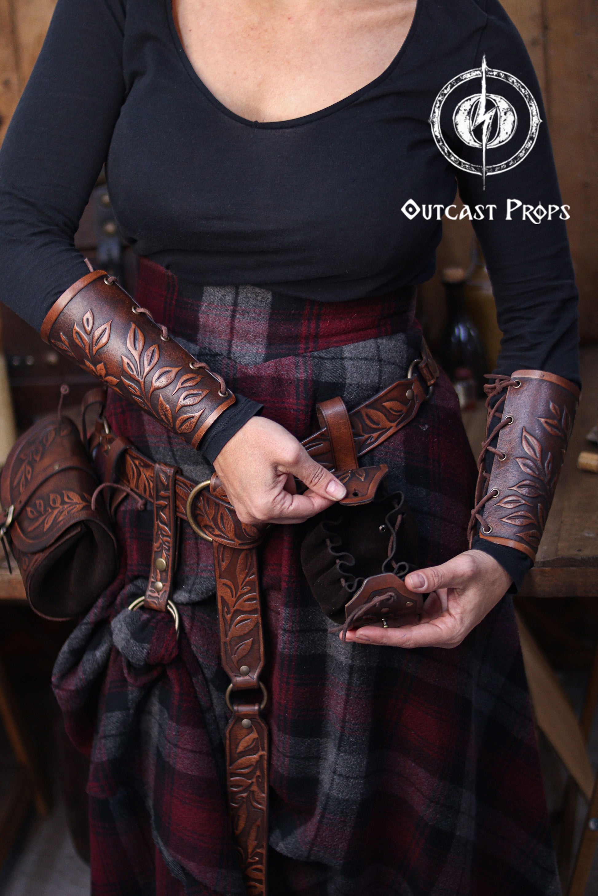 A person wearing brown embossed leather bracers and a matching belt demonstrates how to open a floral leather coin pouch. The plaid skirt and rustic workshop background create a renaissance faire atmosphere. The pouch’s suede interior and drawstring are visible as it is held open. This handcrafted renfaire pouch suits elves, witches, herbalists and nobles, working as a leather coin bag or fantasy dice pouch for immersive LARP and cosplay costumes.