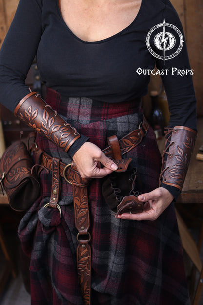 A person wearing brown embossed leather bracers and a matching belt demonstrates how to open a floral leather coin pouch. The plaid skirt and rustic workshop background create a renaissance faire atmosphere. The pouch’s suede interior and drawstring are visible as it is held open. This handcrafted renfaire pouch suits elves, witches, herbalists and nobles, working as a leather coin bag or fantasy dice pouch for immersive LARP and cosplay costumes.