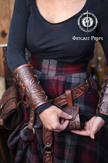 Brown floral leather coin pouch shown in hand as part of a Renaissance Faire herbalist or witch costume, made from vegetable tanned leather and suede. The embossed leaf pattern matches the bracers and belt, ideal for elven characters or nobles who need a stylish pouch for LARP or renfaire. The image shows how the pouch opens with the internal suede bag expanding for coins or trinkets, keeping the elegant fantasy aesthetic intact.