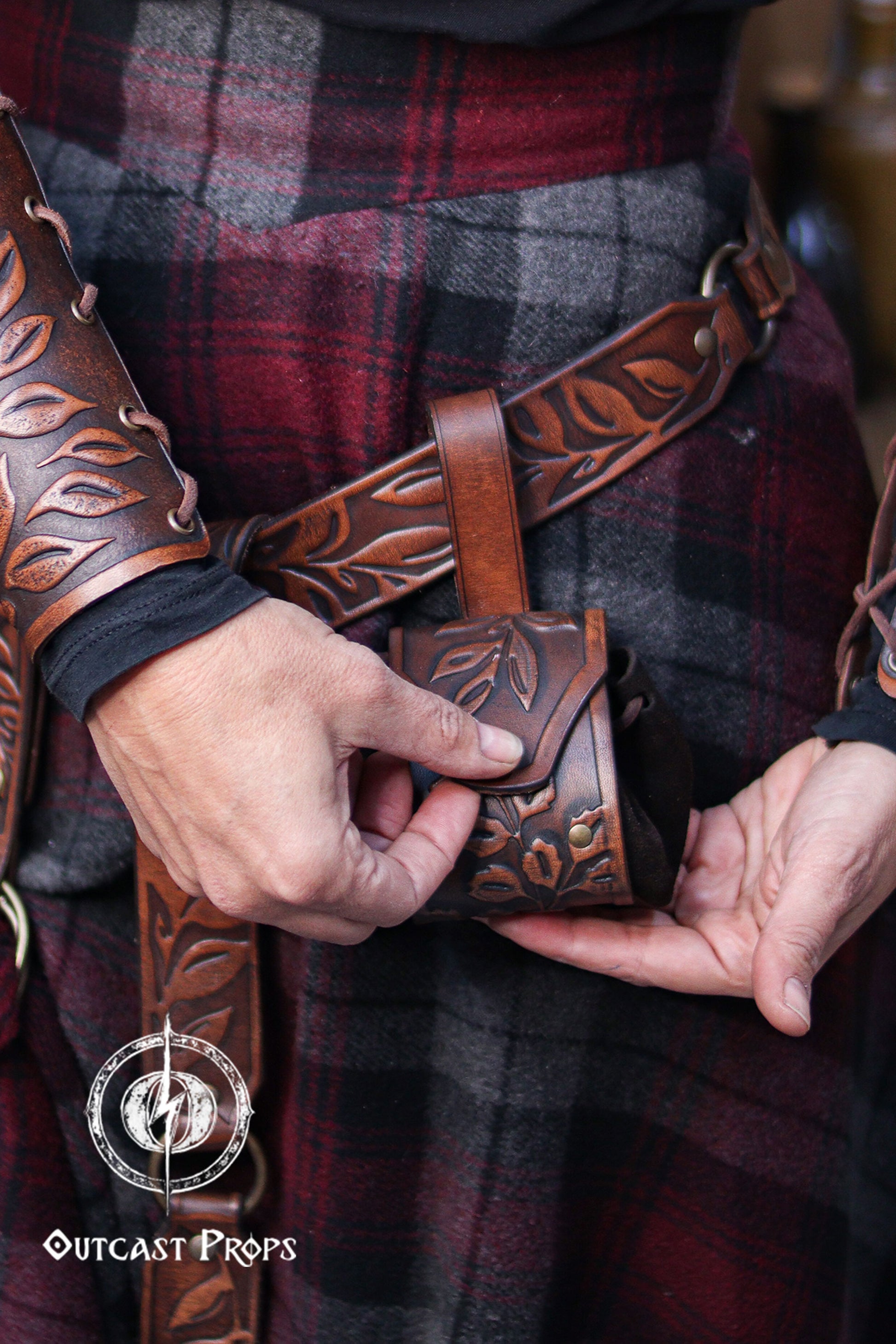 Close view of a person fastening a brown floral embossed leather coin pouch to a matching belt. The handcrafted botanical details and brass rivets stand out against the plaid skirt. The suede body expands to hold coins, dice or small items. Ideal for renaissance faire or renfaire costumes, this fantasy pouch fits elven characters, witches, herbalists and nobles seeking a functional and elegant leather accessory for LARP or cosplay events.