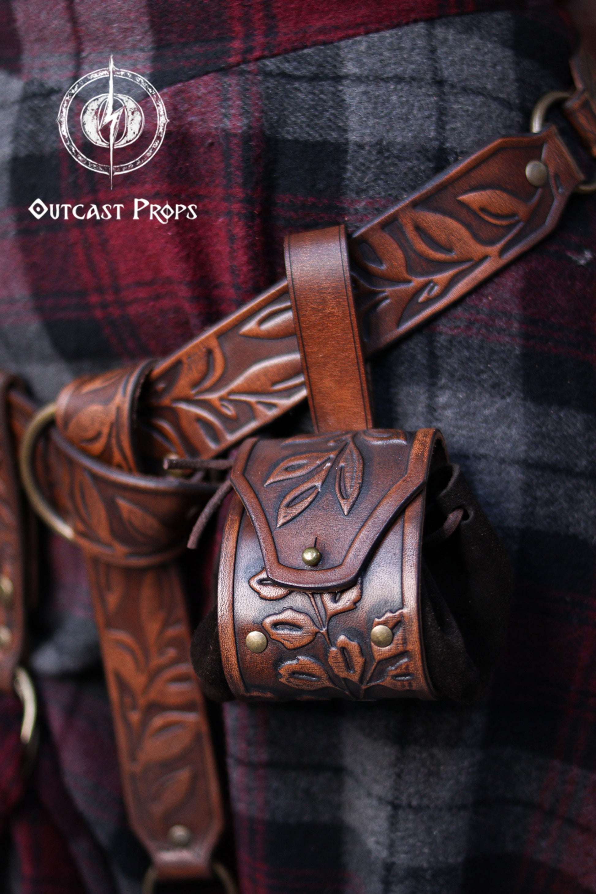 A brown floral embossed leather coin pouch hangs from a matching carved belt worn over a red and grey plaid skirt. The botanical design and aged leather finish give it a natural elven or druidic style. Perfect for renaissance faire or renfaire outfits, the pouch offers a soft suede body for carrying coins, dice or tiny essentials. A practical accessory for witches, herbalists, nobles or fantasy LARP characters seeking detailed leather gear.