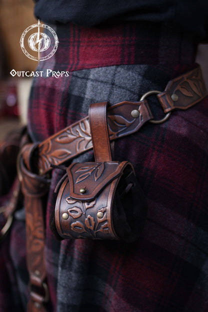 A brown embossed leather coin pouch hangs from a matching belt with botanical carvings. The pouch’s floral flap and suede sides match the natural aesthetic of elven, herbalist or witch characters. The plaid fabric adds a renaissance faire feel. This renfaire pouch is ideal for holding coins, dice or small objects during LARP or cosplay events, offering a practical and elegant accessory for nobles and fantasy adventurers.