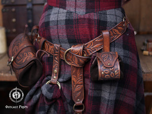 Brown floral leather coin pouch shown in hand as part of a Renaissance Faire herbalist or witch costume, made from vegetable tanned leather and suede. The embossed leaf pattern matches the bracers and belt, ideal for elven characters or nobles who need a stylish pouch for LARP or renfaire. The image shows how the pouch opens with the internal suede bag expanding for coins or trinkets, keeping the elegant fantasy aesthetic intact.