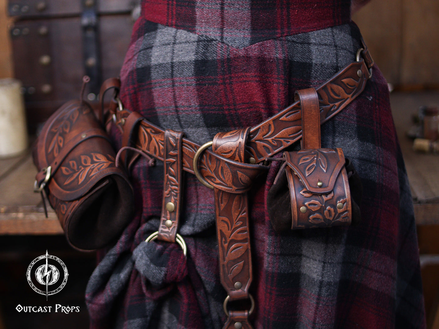 A full brown leather set with embossed botanical patterns is displayed on a person wearing a plaid skirt. The belt carries two accessories: a hipbag and a floral coin pouch. Brass rings and rivets complement the aged leather look. This renaissance faire inspired ensemble suits elves, witches, herbalists and nobles. The coin pouch works as a fantasy dice pouch or coin bag, offering practical storage for LARP and immersive costume wear.