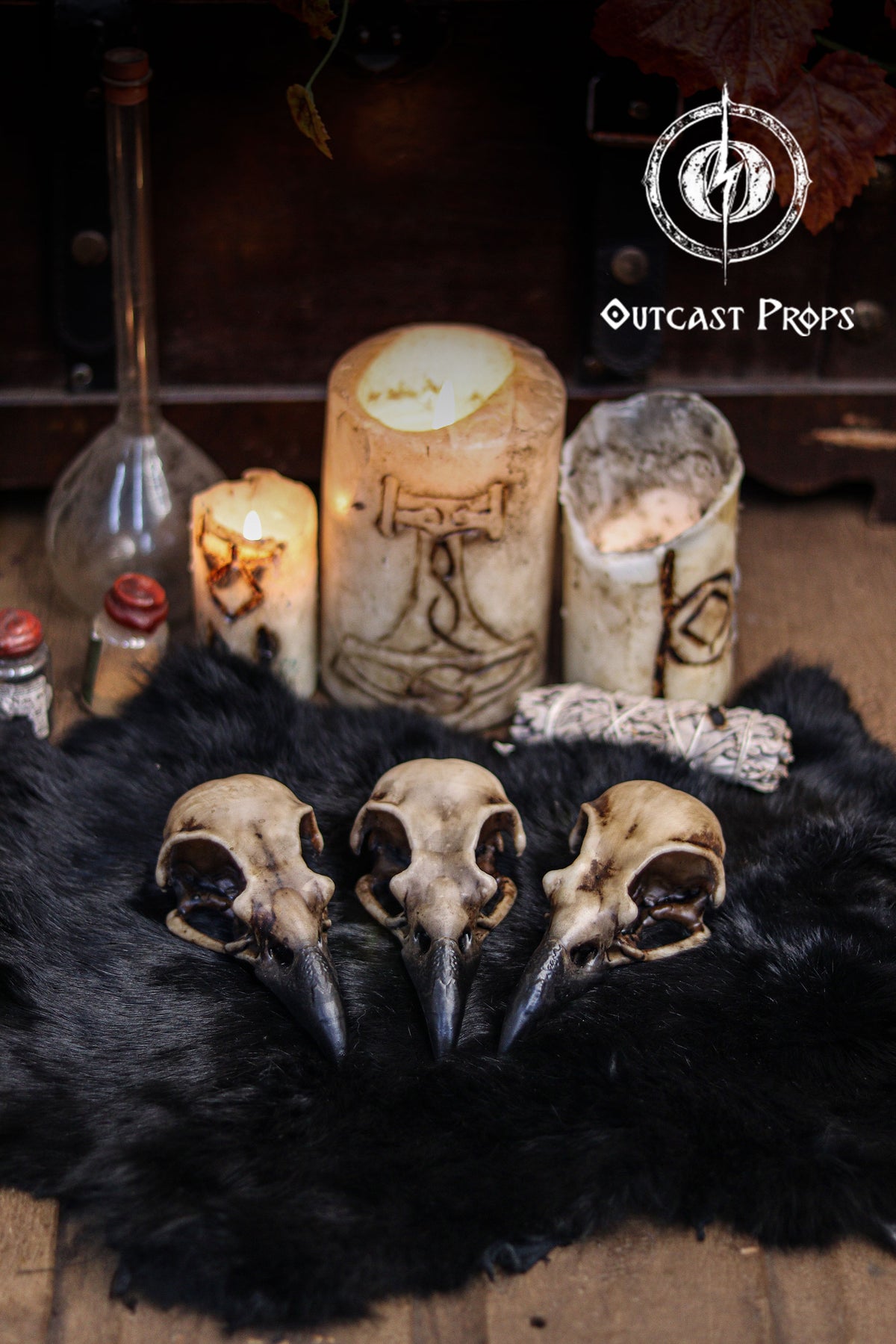 Three resin crow skull replicas painted in aged bone tones rest on a black fur surface, surrounded by gothic decor elements. Behind them are large carved candles, tiny potion bottles and a smudge bundle, creating an atmospheric witchcraft altar scene. The faux bone raven skull replicas show realistic textures and dark beaks, ideal for oddities decor, faux taxidermy displays, jewelry craft, LARP props and cosplay accessories.