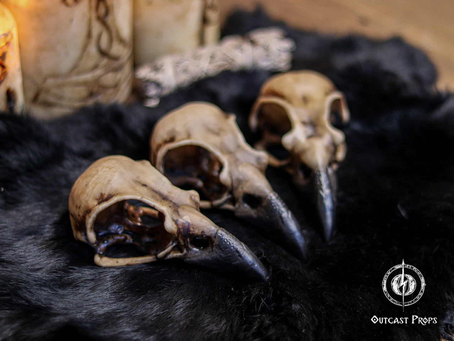 Three faux bone resin crow skull replicas lie on black fur, angled toward the viewer to show detailed texture, weathered bone coloration and hand painted aging. Gothic candles and ritual elements appear softly in the background, setting a witchcraft inspired mood. Ideal for faux taxidermy collectors, gothic decor lovers, oddities decor shelves, jewelry craft supplies, headdress building, cosplay props and LARP accessories requiring realistic raven skull replicas.