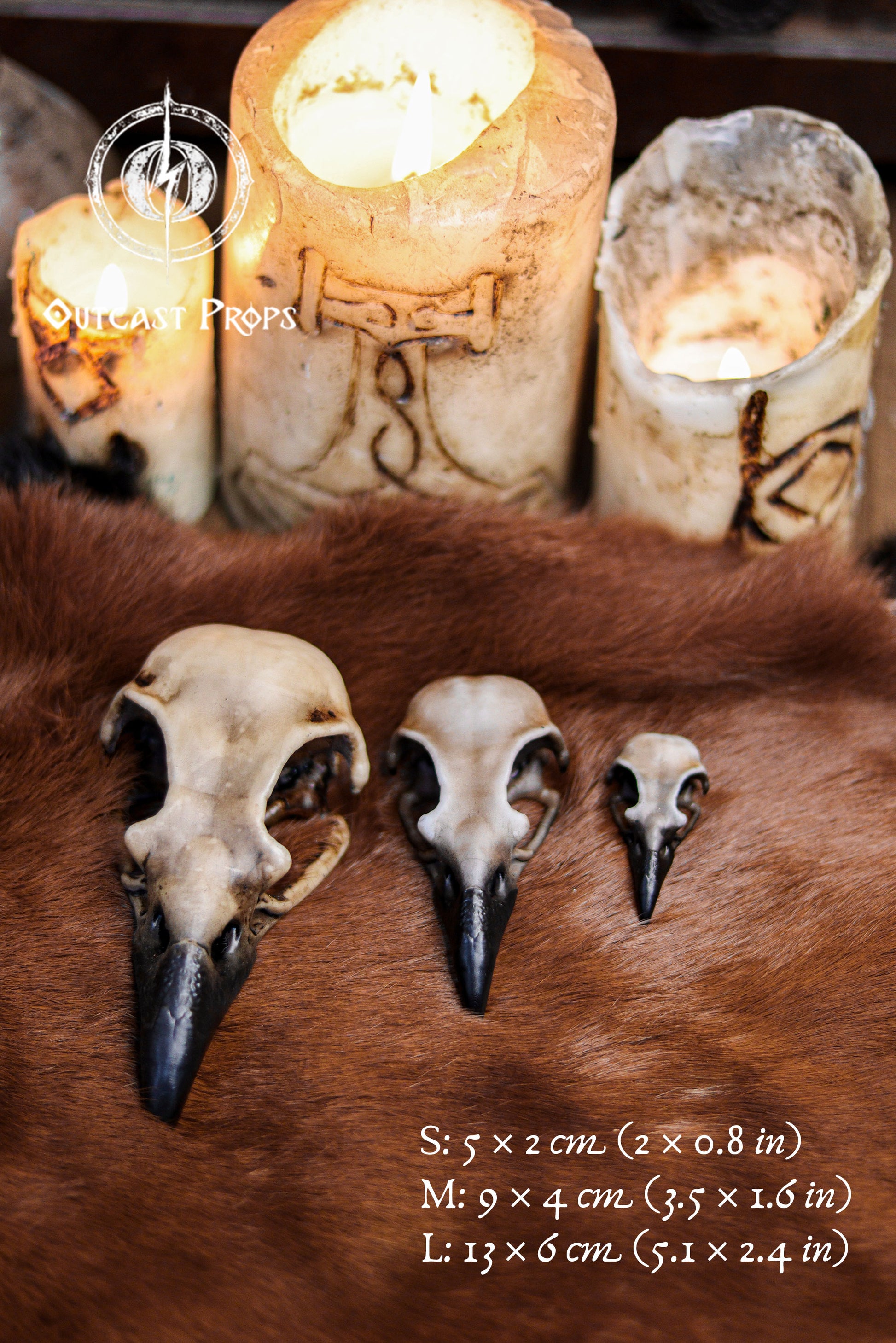 Three resin crow skull replicas in sizes S, M and L are displayed on a brown fur surface with carved candles glowing in the background. The faux bone raven skull replicas show detailed hand painted aged bone texture and dark beaks. Size measurements are written on the image for clarity. This atmospheric setup evokes gothic decor, oddities decor and faux taxidermy art, perfect for jewelry craft components, cosplay props and LARP accessories.