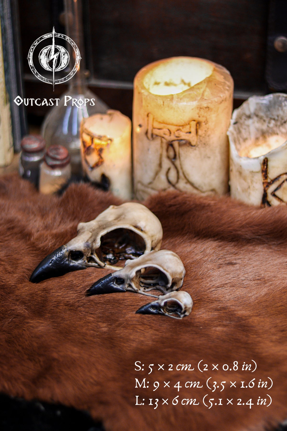Three resin crow skull replicas in sizes S, M and L rest on brown fur beside carved candles and vintage bottles, creating a witchcraft inspired setting. The faux bone raven skull replicas display realistic aged bone shading and dark beaks. Text on the image lists measurements for each size. Ideal for gothic decor, oddities decor collectors, faux taxidermy displays, jewelry craft supplies, cosplay builds and detailed LARP props.