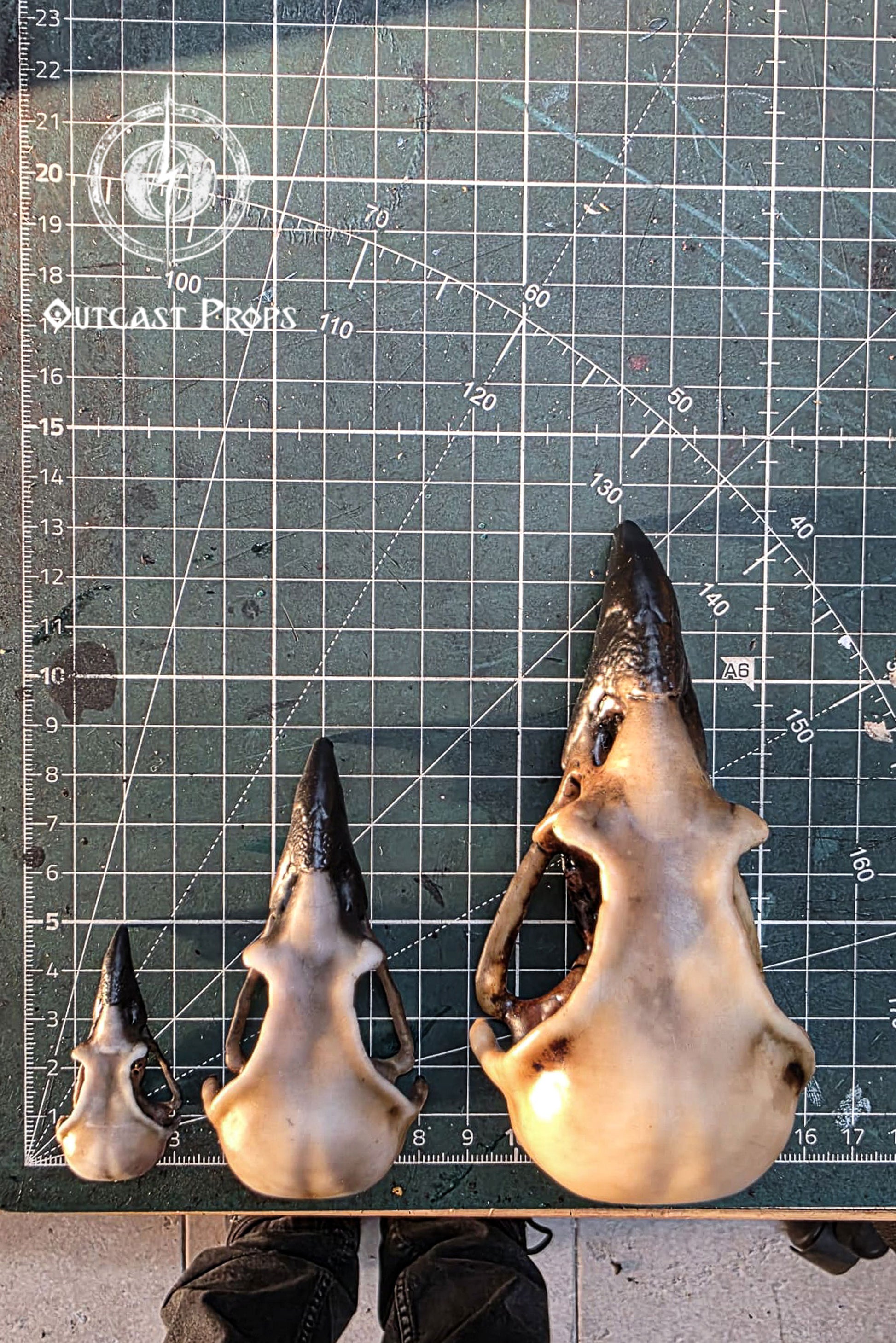 Three resin crow skull replicas in sizes S, M and L are displayed vertically on a large cutting mat with measurement grid, showing their scale. The faux bone raven skull replicas feature hand painted aged bone texture and dark beaks. The grid background helps viewers compare proportions for jewelry craft, cosplay builds, gothic decor, oddities decor, faux taxidermy displays and LARP props where accurate size reference is important.