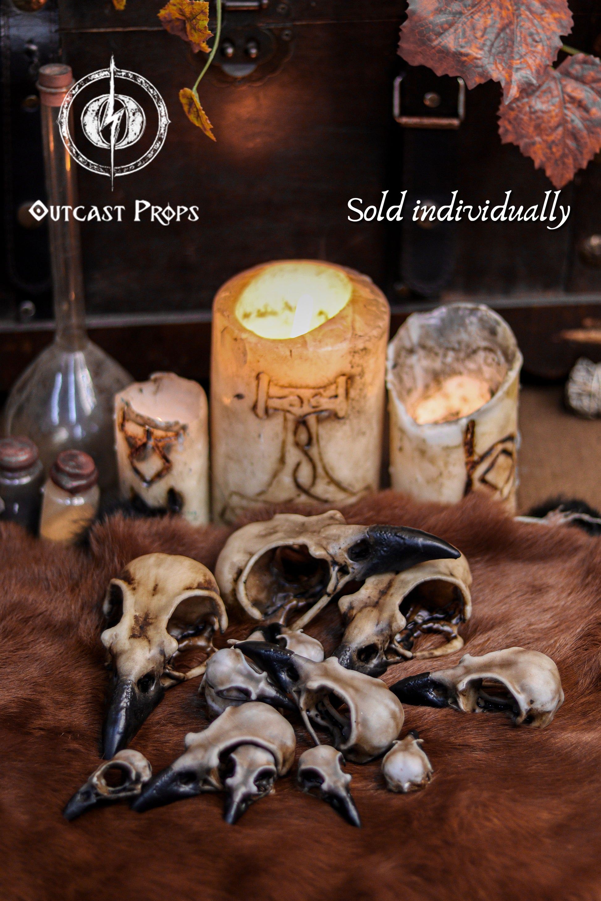 A mixed collection of resin crow skull replicas in various sizes is arranged on brown fur in front of carved candles and potion bottles, with text noting they are sold individually. The faux bone raven skull replicas show aged bone paint, dark beaks and realistic details. Perfect for gothic decor, oddities decor, faux taxidermy sets, jewelry craft, costume making, cosplay props and immersive LARP accessories.