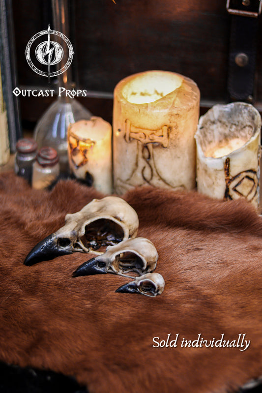 Three resin crow skull replicas in sizes L, M and S are arranged on warm brown fur with carved candles glowing softly in the background. The faux bone raven skull replicas show detailed hand painted aging, hollow interiors and dark beaks. Text on the image reads “3 different sizes”. This display highlights their use for gothic decor, oddities decor, faux taxidermy art, jewelry craft components, cosplay props and immersive LARP accessories.