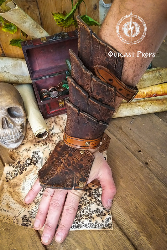 Close-up of a brown leather dragon scale bracer worn on a male hand, displayed over a vintage map with coins, a skull, and scrolls. This handmade bracer is ideal for LARP, pirate cosplay, monster hunters, or fantasy warriors. Crafted from premium vegetable-tanned leather and finished with Outcast Props' signature aging techniques, each piece is unique and tells a story straight from an epic adventure.