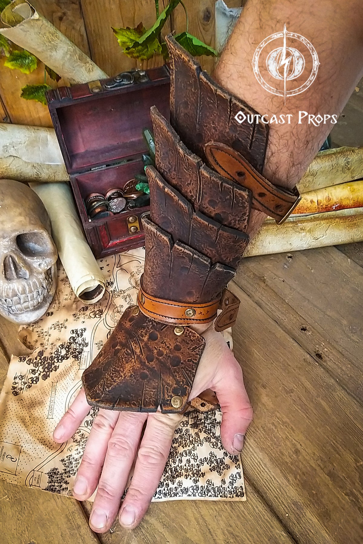 Close-up of a brown leather dragon scale bracer worn on a male hand, displayed over a vintage map with coins, a skull, and scrolls. This handmade bracer is ideal for LARP, pirate cosplay, monster hunters, or fantasy warriors. Crafted from premium vegetable-tanned leather and finished with Outcast Props' signature aging techniques, each piece is unique and tells a story straight from an epic adventure.