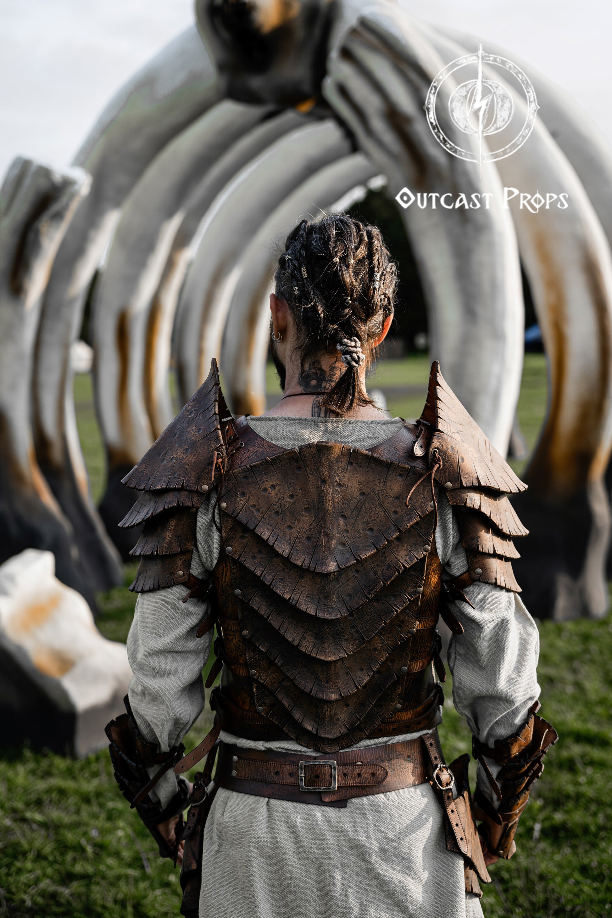 Back view of a warrior wearing the Dragon Scales Armor Set, standing before a giant bone-like sculpture at a LARP event. The armor is handmade from premium vegetable-tanned leather, textured to mimic ancient dragon hide. Shoulder guards and layered plates give a fierce, battle-ready look, ideal for cosplay, fantasy photoshoots, and immersive roleplay at Ren Faires or Viking festivals.