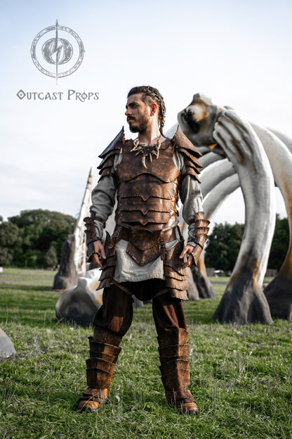 Full body photo of a LARP player dressed in the Dragon Scales Armor Set, in front of monumental bone structures. The complete leather outfit includes breastplate, pauldrons, arm guards, tassets, belt and greaves, all textured and dyed to resemble dragon hide. This fantasy armor is perfect for epic cosplay, Viking events, or fantasy reenactments. Made from eco-friendly leather and finished with Outcast Props’ signature techniques.