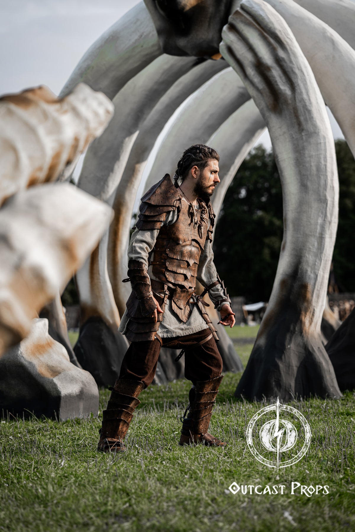 Full body view of a fantasy warrior wearing the Dragon Scales armor set by Outcast Props. Posing in a LARP environment under large skeletal structures, this armor is built with layered leather scales and rugged linen. Ideal for renaissance fairs, fantasy LARP and immersive cosplay. Crafted from vegetable-tanned leather with natural dyes, blending style and comfort.