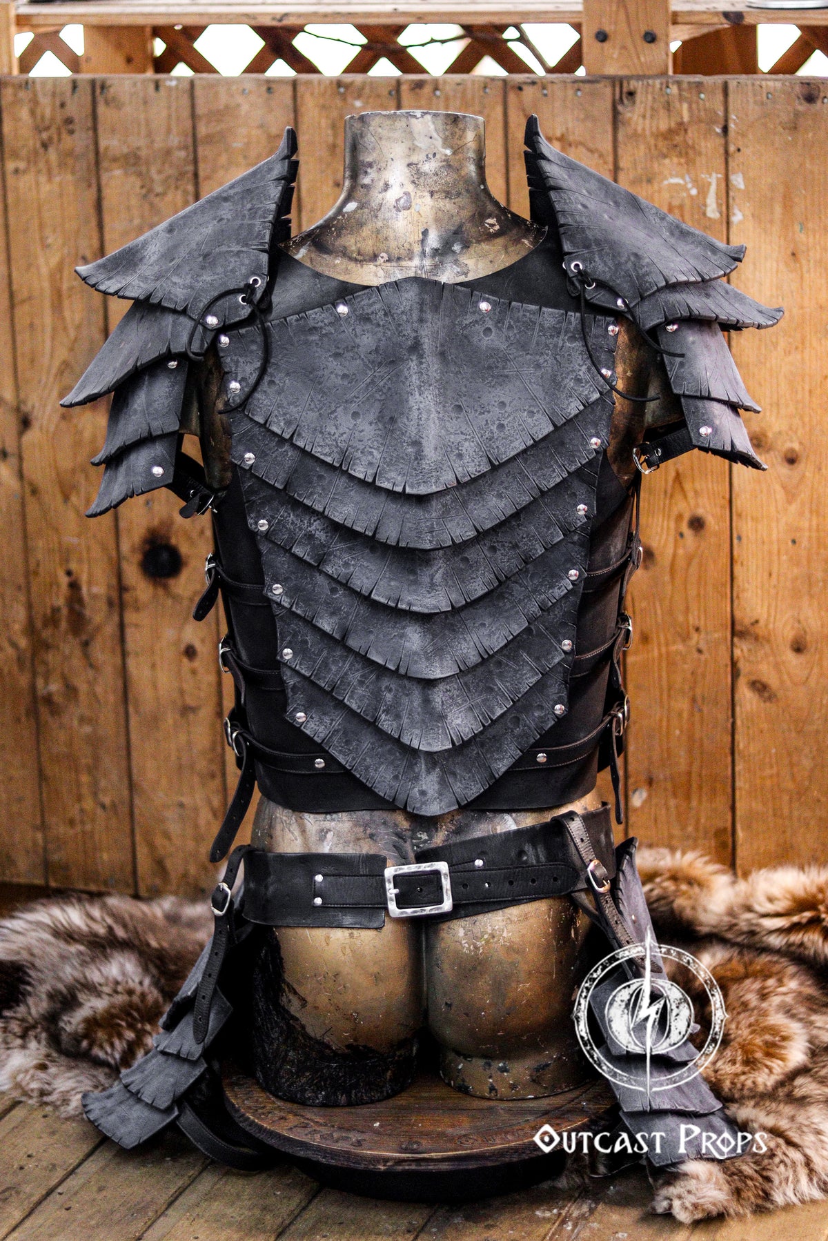 Rear view of black dragon scale leather armor set displayed on a vintage mannequin. Includes shoulder pauldrons, layered chestplate, side buckles, and matching belt. The deep textures and weathered finish give it a dark fantasy style, suitable for LARP warriors, mercenaries, or dark elves. Perfect for gothic or villainous characters at Renaissance fairs or immersive fantasy events. Crafted by hand with Outcast Props’ unique leather aging techniques. Medieval armor on a mannequin against a wooden background