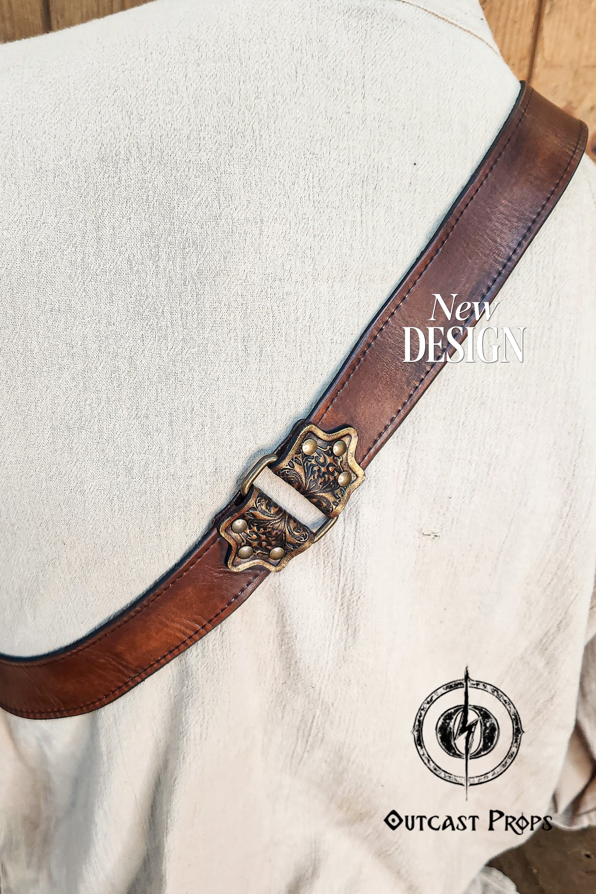Close-up of a handmade leather baldric strap resting diagonally across the back of a cream medieval tunic. The strap features premium brown vegetable-tanned leather and an ornate brass buckle with floral motifs. Ideal for pirate cosplay, steampunk outfits, or LARP characters. The detailed stitching and leather aging add to its realism and craftsmanship. Perfect accessory for historical reenactments or fantasy events.