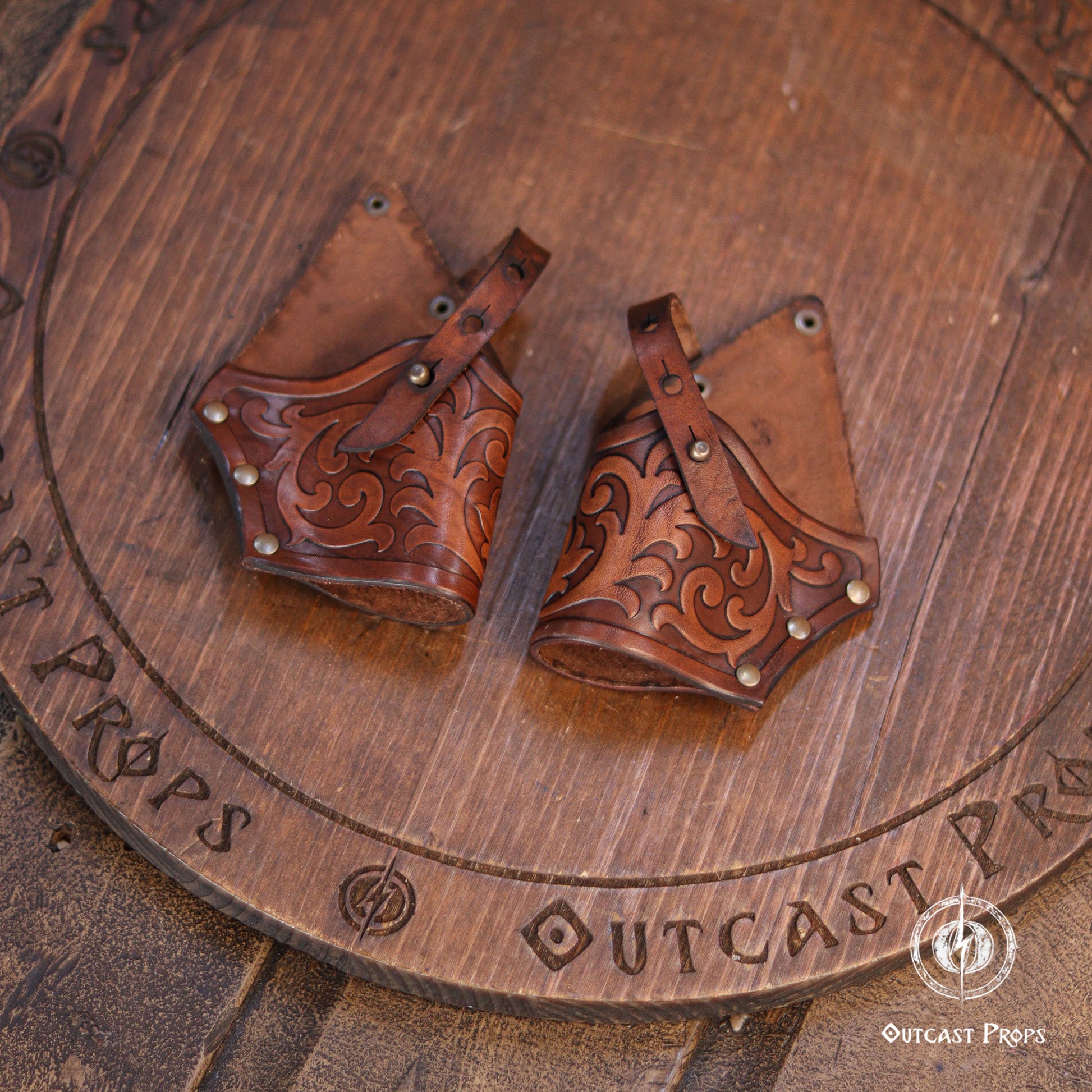 A pair of brown leather prop sheaths with intricate hand-tooled floral carvings, displayed on a rustic wooden surface engraved with 'OUTCAST PROPS'.  A pair of brown leather flintlock holsters featuring intricate embossed scrollwork patterns, secured with adjustable straps and metallic rivets. The leather has a polished finish with darker stained details highlighting the carved designs.