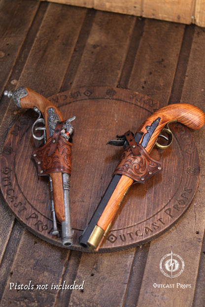 Two ornate replica flintlock pistols with carved brown leather sheaths and wooden grips, displayed on a round engraved wooden board over a rustic wood plank surface. A watermark reads 'Pistols not included' and 'Outcast Props' appears at the bottom right. Two brown leather holsters with intricate embossed scrollwork patterns, each designed to hold a pistol. The holsters are displayed on a circular wooden board featuring the engraved text OUTCAST PROPS. Pistols not included.