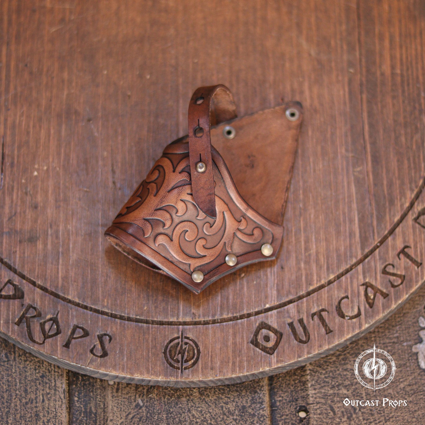 Brown leather holster featuring intricate embossed scrollwork patterns, a vertical strap with metal rivets, and a triangular shape. The leather has a polished finish with darker shading in the recessed areas of the decorative carvings.