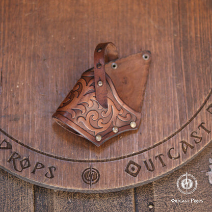 Brown leather holster featuring intricate embossed scrollwork patterns, a vertical strap with metal rivets, and a triangular shape. The leather has a polished finish with darker shading in the recessed areas of the decorative carvings.