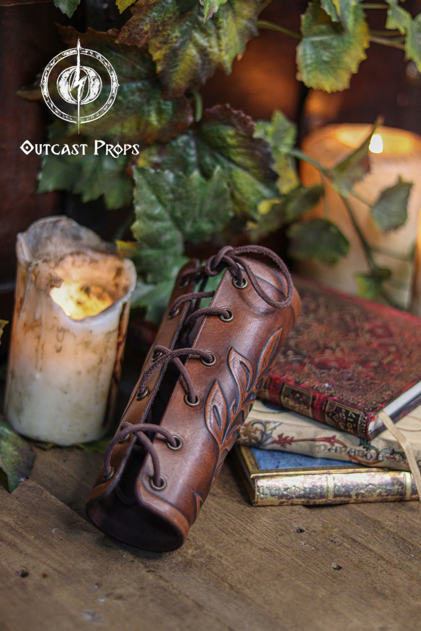 A brown leaf elven bracer made of premium vegetable tanned leather lies beside old books, a melted candle and green foliage. Its embossed leaf motif and laced closure create an elegant renaissance faire accessory ideal for elves, witches, herbalists, nobles and fae costumes. This leather larp bracer also fits roman laurel or Caesar cosplay thanks to its classic leaf design. Warm lighting highlights the hand aged details and fantasy aesthetic.