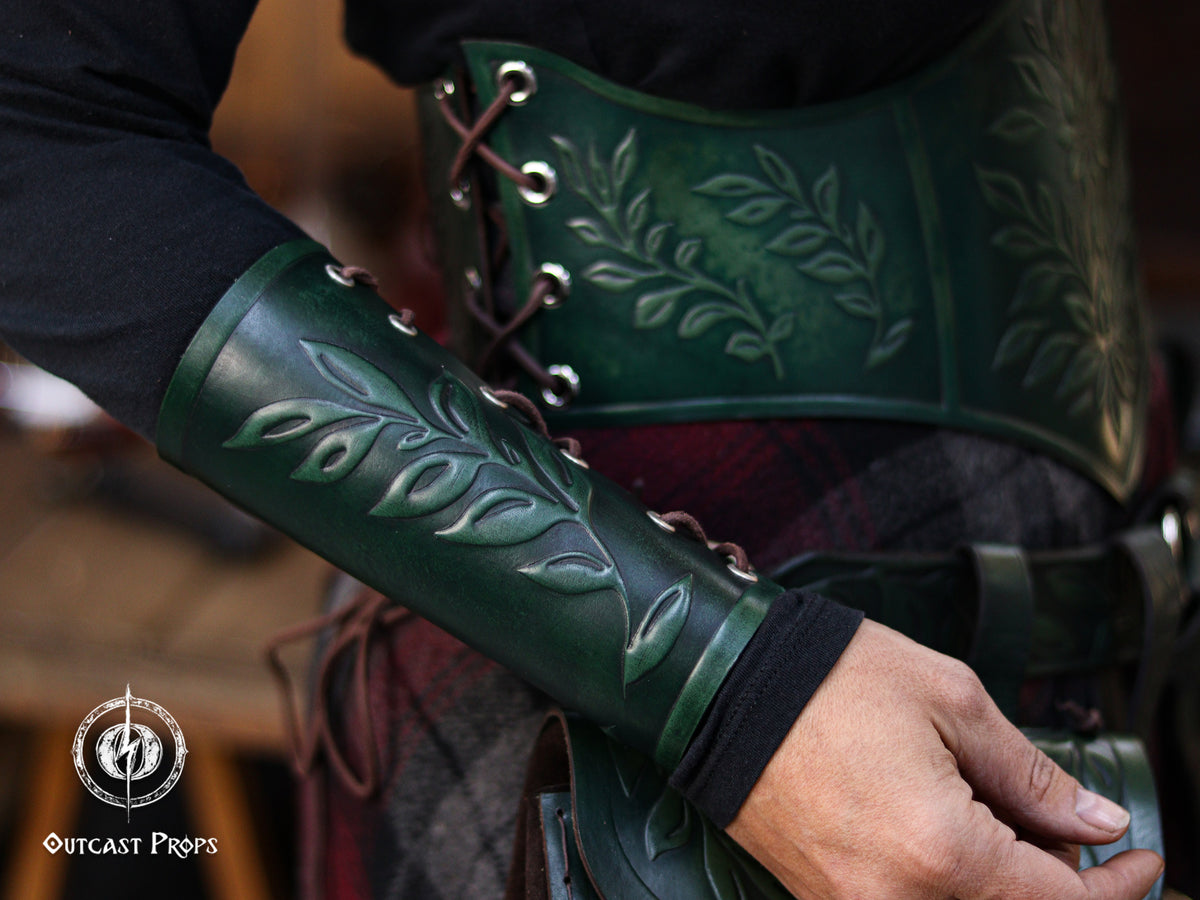 Close up of a green leaf elven bracer worn on the forearm with a matching leather hip bag featuring embossed leaf designs. Perfect for renaissance faire costumes, elves, witches, herbalists, fae characters and larp outfits. The elegant leaf motif also works for roman laurel or Caesar cosplay. The rustic background enhances the handcrafted aged leather and natural fantasy aesthetic of this coordinated accessory set.