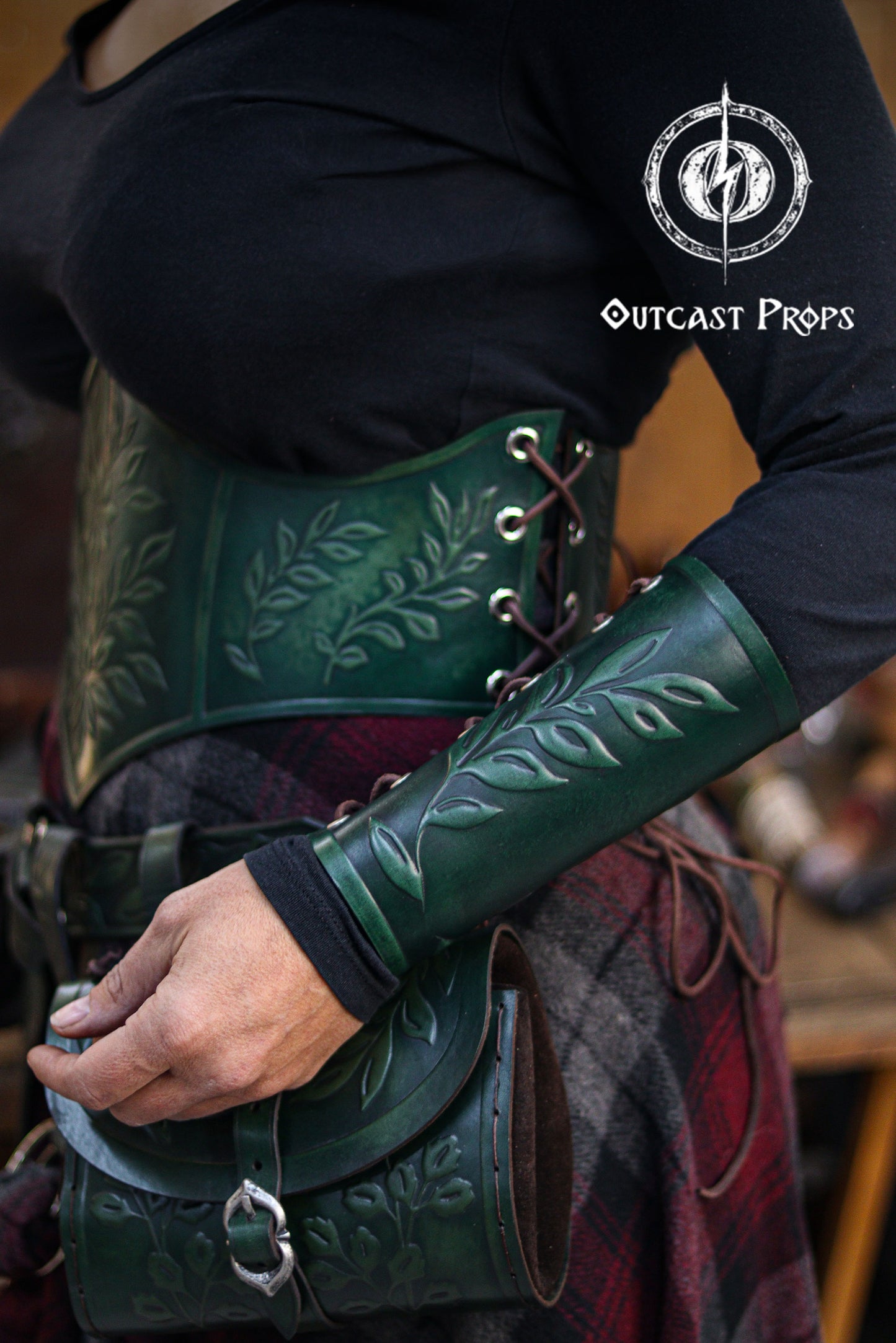 Side view of a woman wearing a green leaf elven bracer with a matching corset and hip bag decorated with embossed leaf patterns. Ideal for renaissance faire fashion, larp characters, elves, witches, herbalists, nobles and fae performers. The leaf motif also suits roman laurel or Caesar themed cosplay. Soft lighting and rustic textures highlight the hand aged leather and elegant fantasy craftsmanship of the ensemble.