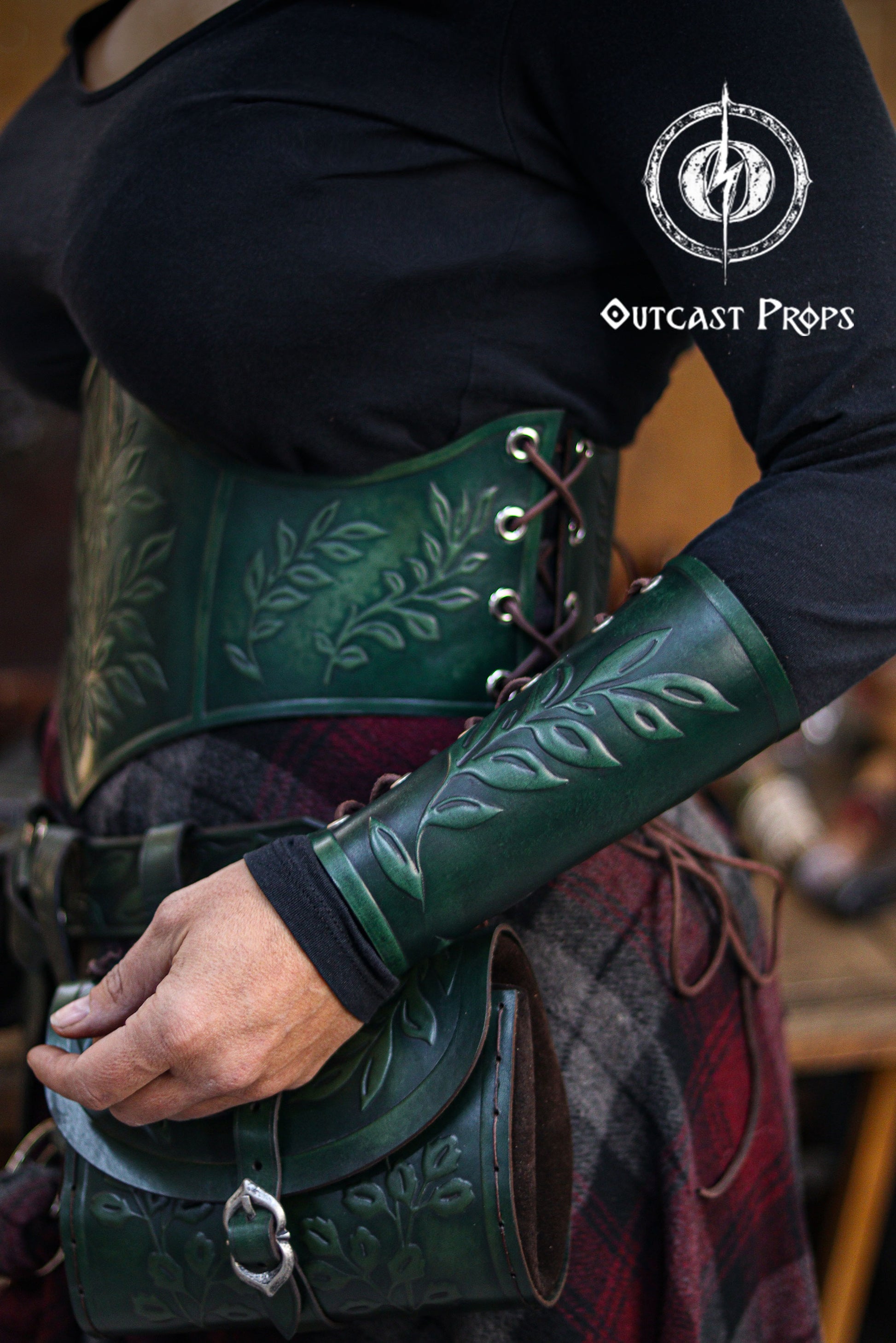 Side view of a woman wearing a green leaf elven bracer with a matching corset and hip bag decorated with embossed leaf patterns. Ideal for renaissance faire fashion, larp characters, elves, witches, herbalists, nobles and fae performers. The leaf motif also suits roman laurel or Caesar themed cosplay. Soft lighting and rustic textures highlight the hand aged leather and elegant fantasy craftsmanship of the ensemble.